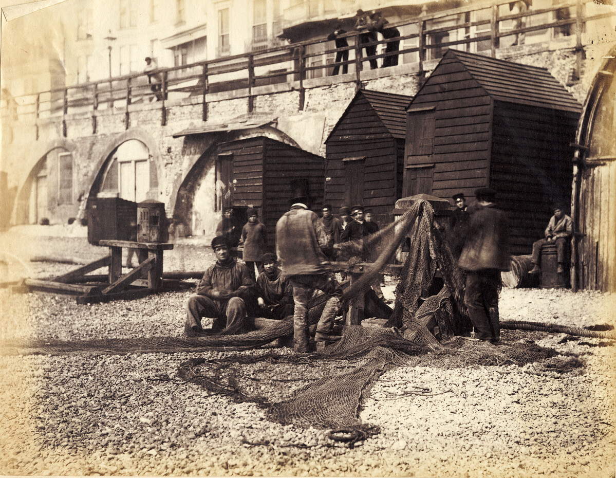 Sepia photograph showing fishermen on Brighton beach with a net and a hut in the background.