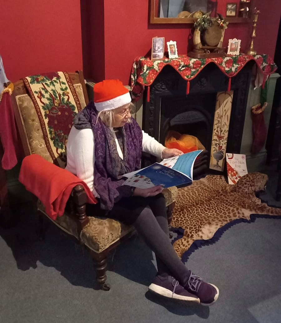 A woman sits on a chair reading a book. She's wearing a santa hat.