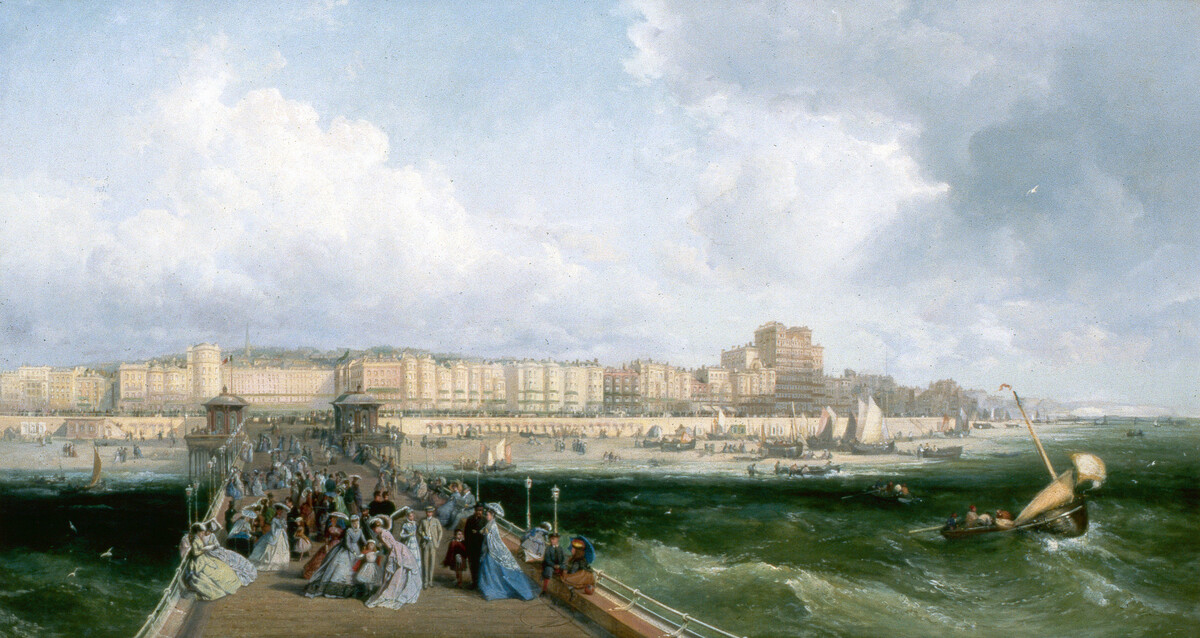 The West Pier, Brighton, is seen from about half way along it, looking landwards. The pier is thronged with figures in late Victorian dress. The seafront is seen from Preston Street to the Chain Pier at far right. Fishing boats at sea in right foreground