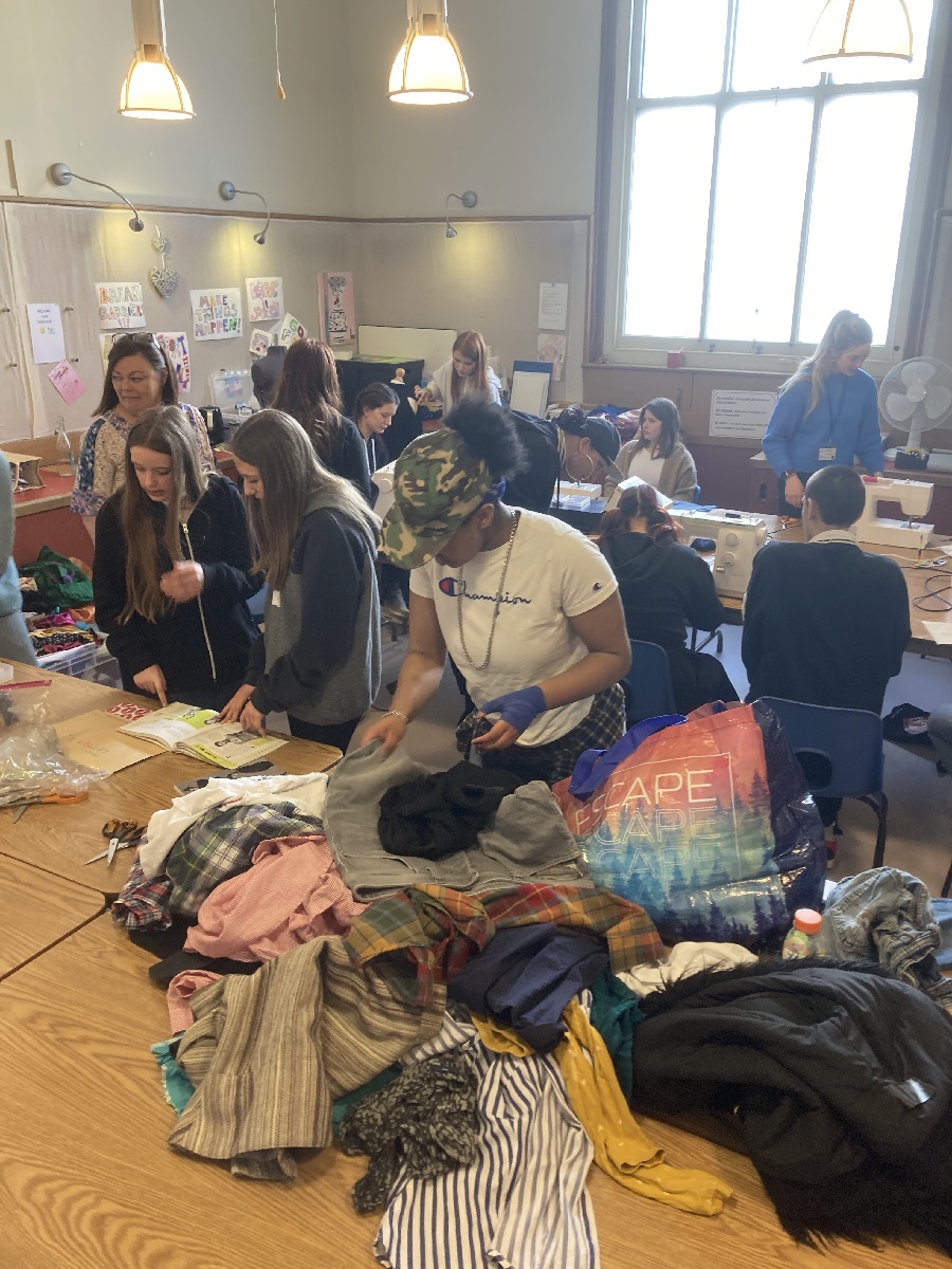 Photograph of the Fast Fashion with the Young Carers event. A group of young people look through a pile of clothes. In the background others are sat by sewing machines