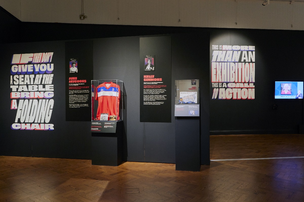 Goal Power! exhibition in Brighton Museum & Art Gallery. There are two football shirts in cases with accompanying text panels on the wall. A screen plays an interview and there are slogans printed on the walls.