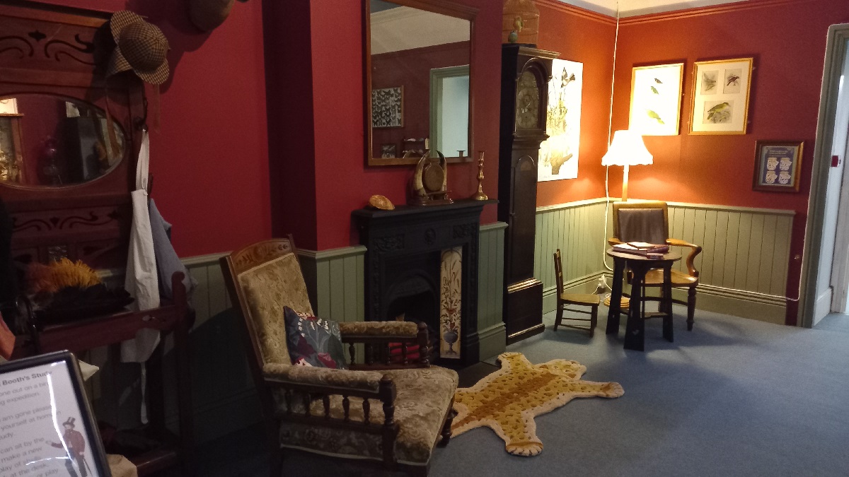 The Victorian Parlour at the Booth Museum. There is a fireplace in the centre with a fake animal skin rug. An armchair is next to it. A desk and chair are in the far corner.