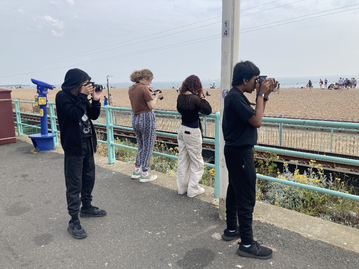 Brighton Photo Club, Rachel Poulton. A group of young people stand along the seafront holding cameras, which point to the beach