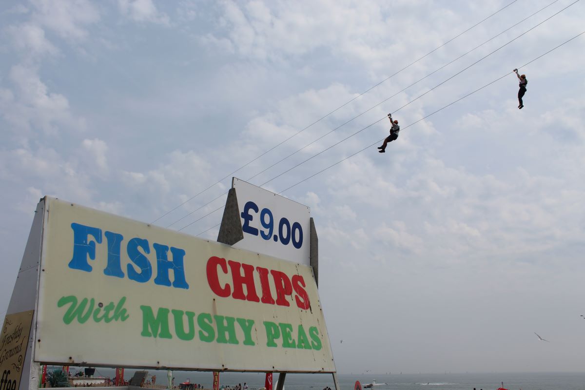In this photo, you can see the Brighton seaside with two people racing down a dual zipline, and there's also a prominent "Fish and Chips" sign.