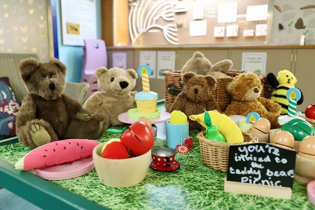A Teddy Bear’s Picnic at the Booth Museum. A group of soft toys sit with toy food and a chalk board sign which reads You're invited to the teddy bear's picnic
