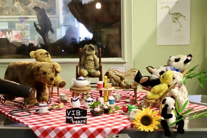 Very Important Bears at A Teddy Bear’s Picnic at the Booth Museum. A group of older soft toy bears from our toy collection sit at a separate picnic with a blackboard sign for V.I.Bs