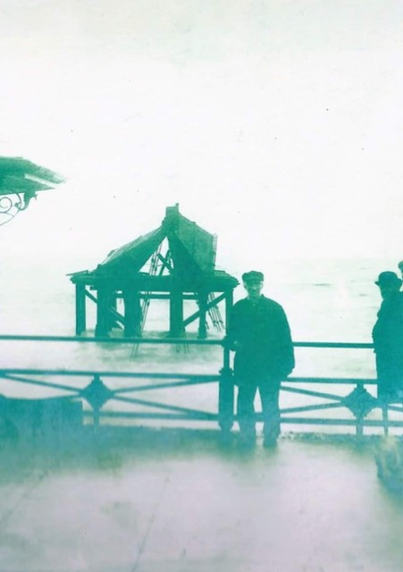 The Last Man on the Chain Pier. An old faded photograph of a man in a cap and long coat stood against seafront railings with the destroyed Chain Pier in the background.