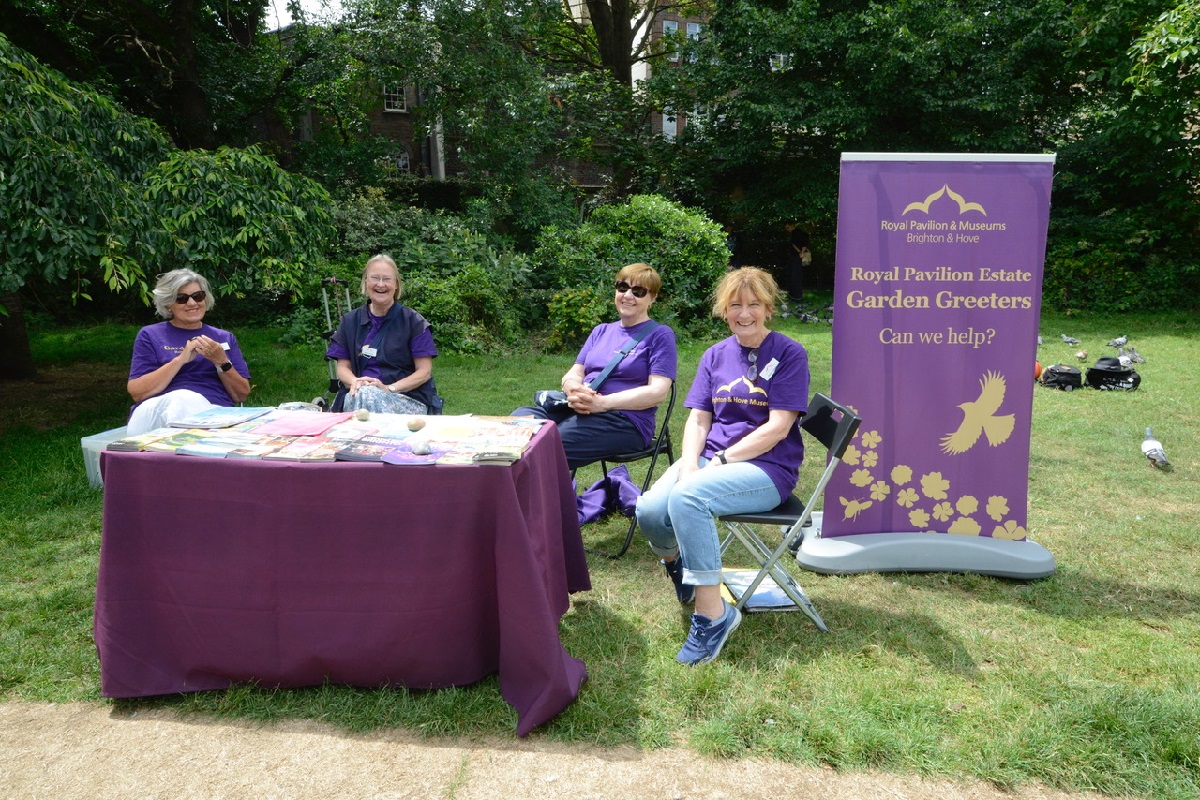 Garden Greeters in the Royal Pavilion Garden. Four women in purple Garden Greeter t-shirts are sitting at a table in the garden. The table is dressed with a purple cloth and information leaflets and a purple Garden Greeters banner stands to the side.