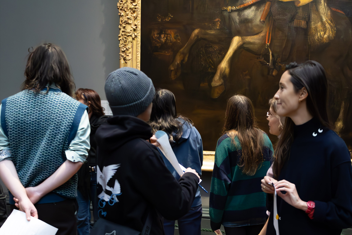 Photography Club's visit to the National Gallery. A group of young people stand in a gallery looking at portraits, a woman talks to a boy as he looks at the portrait