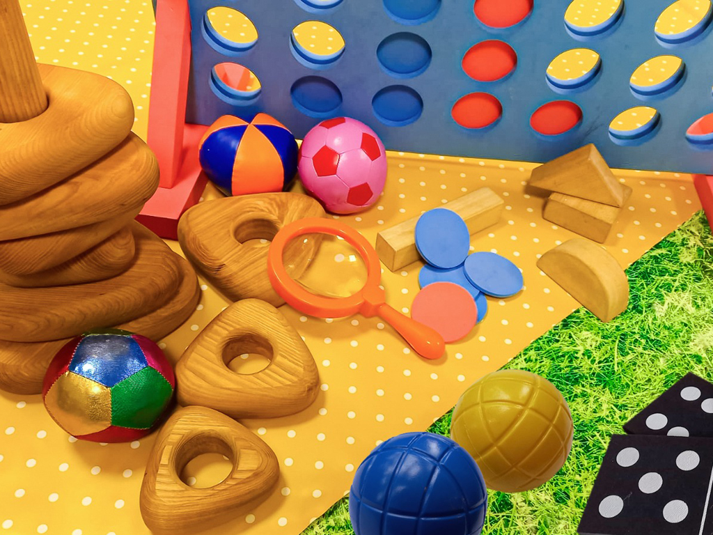 Play days at Hove. A variety of children's toy are displayed on a mat on the grass. Giant Dominos, Connect 4, juggling balls, wooden stacking toys and bat and balls are among them