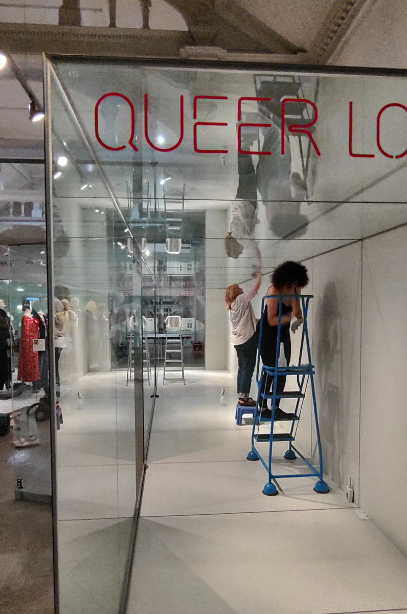 The Collections team clean the case for Queer Looks. The large case is empty of mannequins, two members of the team are inside, one wipes the back of the case, the other is on a step ladder