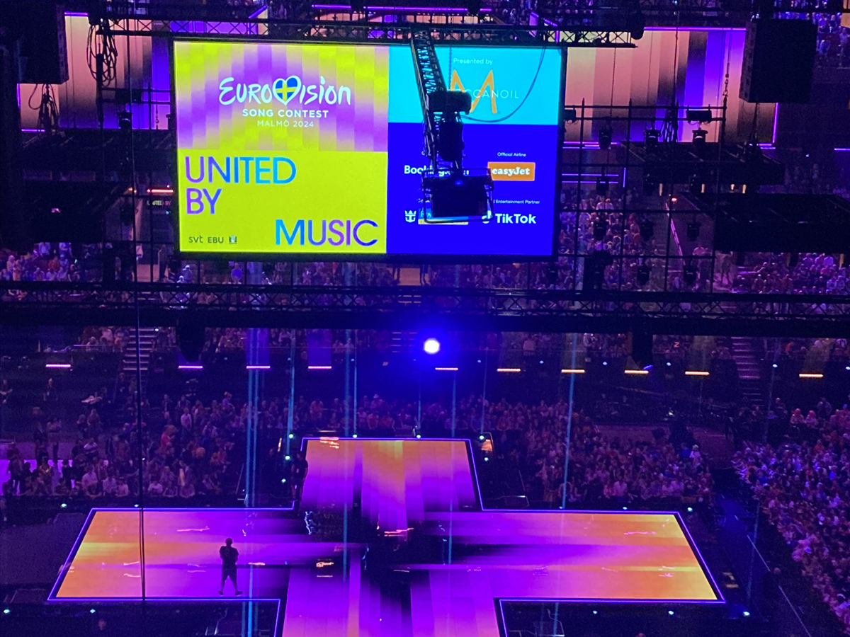 Eurovision final stage in the Arena. The stage is a large cross in the centre of the arena with the crowd around it. Large screens are overhead.