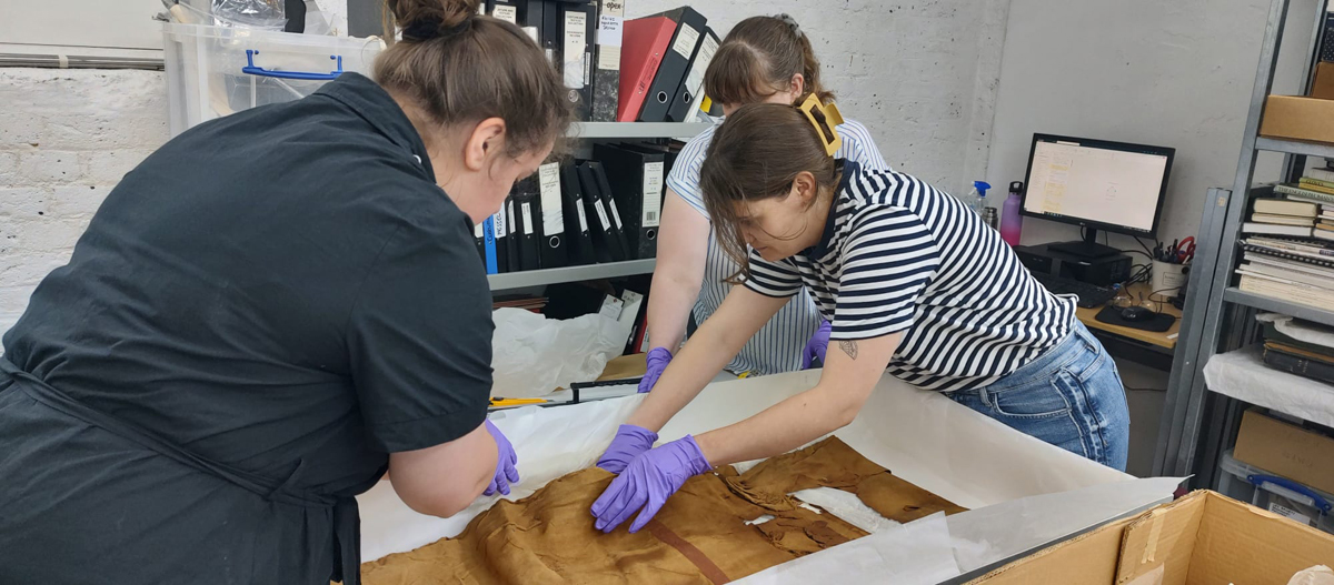 Repacking the World Art textiles in the store. Three women wearing gloves are carefully handling textiles in a large box placed on a table.