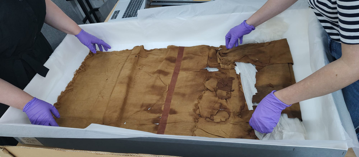 Repacking the World Art textiles in the store. Two sets of gloved hands can be seen carefully touching a textile in a large box.