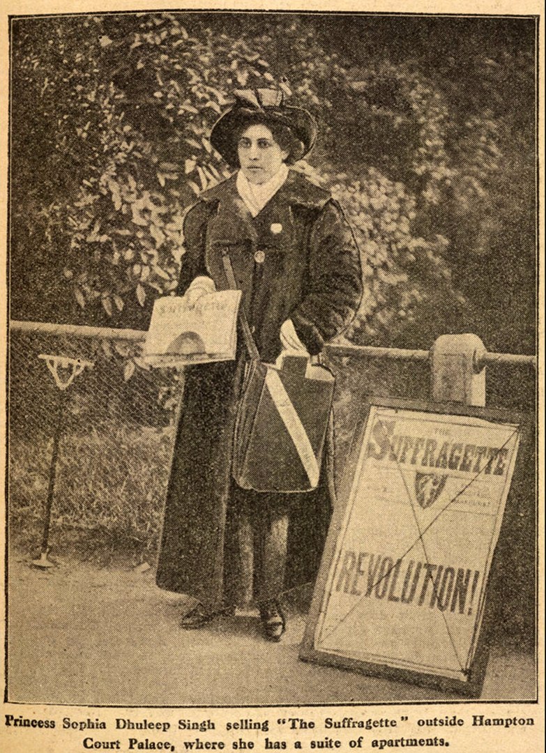 A photograph of an Indian woman, wearing a full-length coat, hat with a cockade, with a large satchel slung across her body, holding a copy of The Suffragette newspaper in her right hand next to an advertising board for the paper proclaiming REVOLUTION!