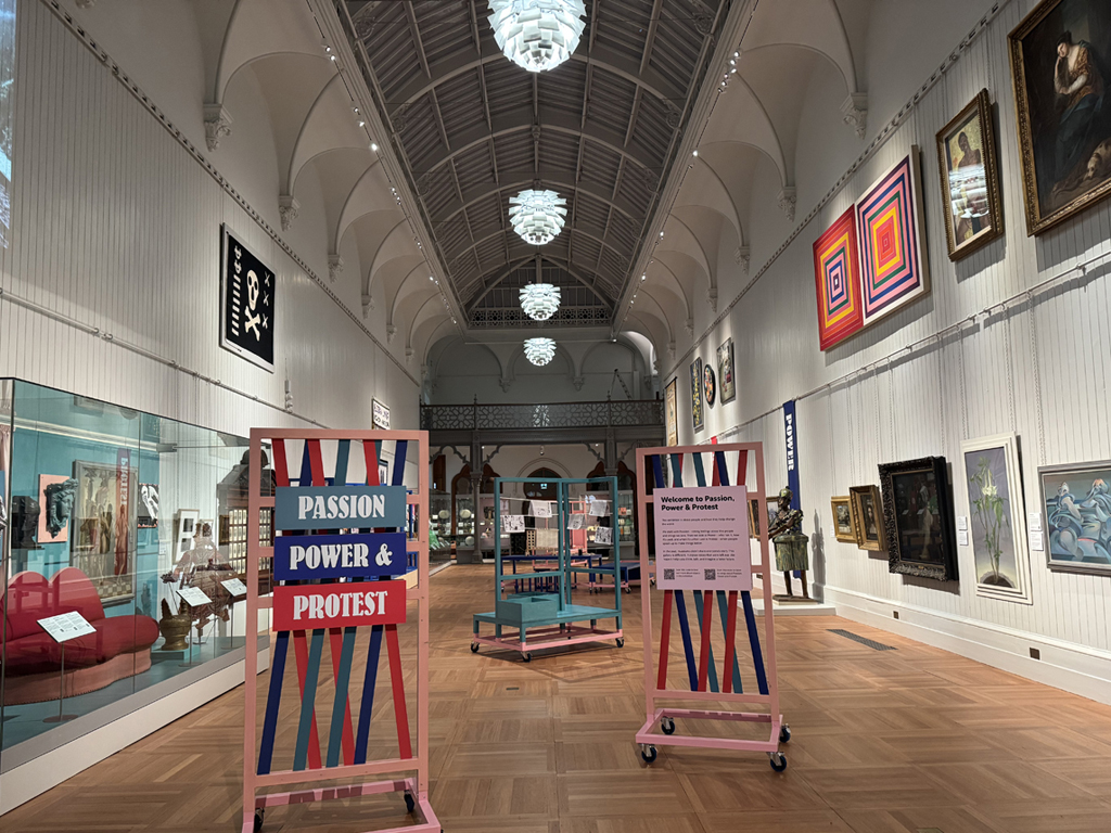 Centrall Gallery of Brighton Museum & Art Gallery. Art works line the wall on the right, cases of objects can be seen on the left. Moveable signs and tables are in the centre