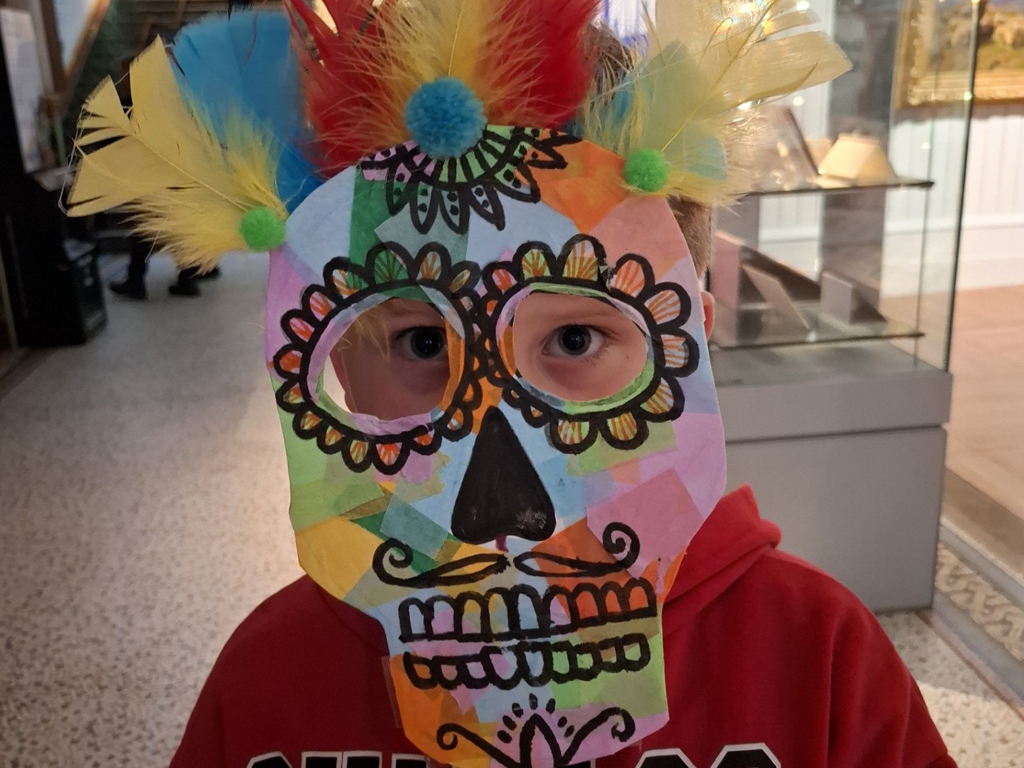 Art Club at Brighton Museum. A boy holds a mask to his face. The mask is a skull with feathers attached to the top.