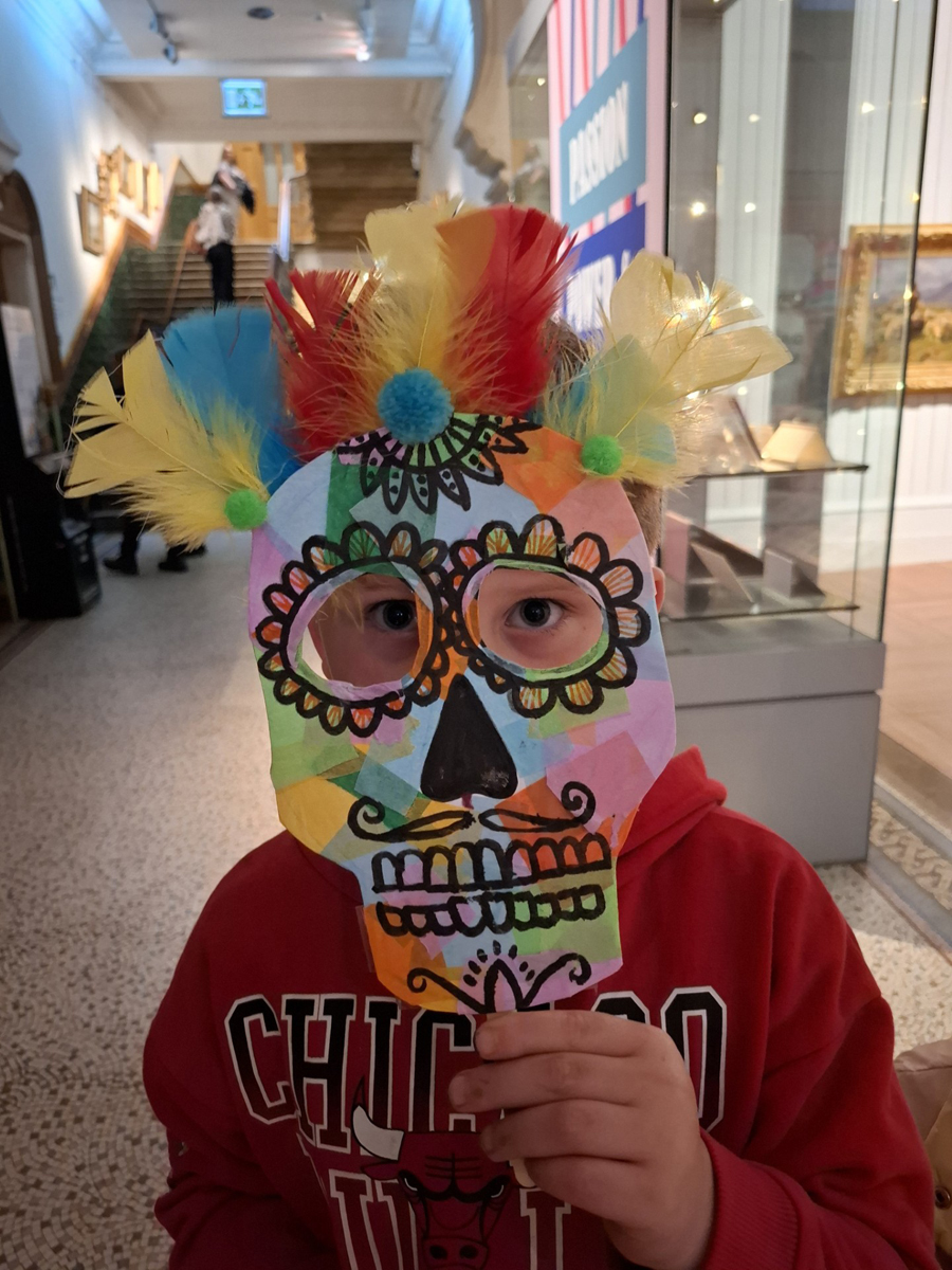 Art Club at Brighton Museum. A boy holds a mask to his face. The mask is a skull with feathers attached to the top.