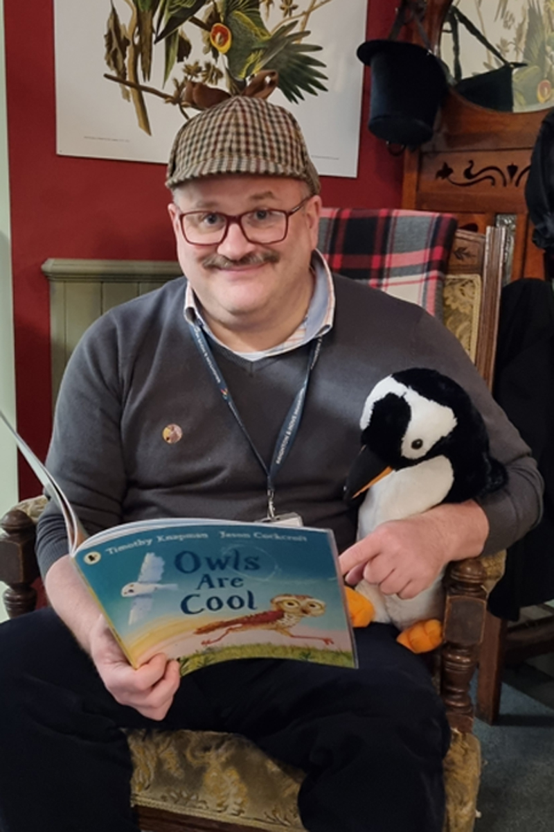 Storytelling at Brighton Museum. A smiling man sits in a chair holding a children's book, he has a toy stuffed penguin under his arm