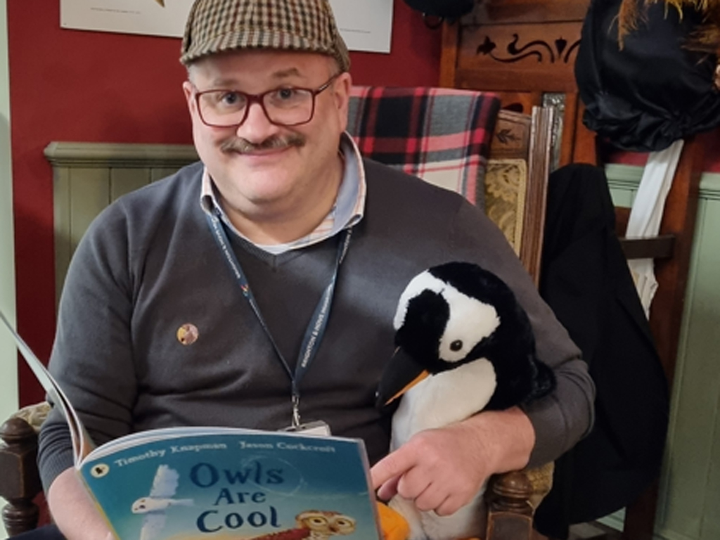 Storytelling at Brighton Museum. A smiling man sits in a chair holding a children's book, he has a toy stuffed penguin under his arm