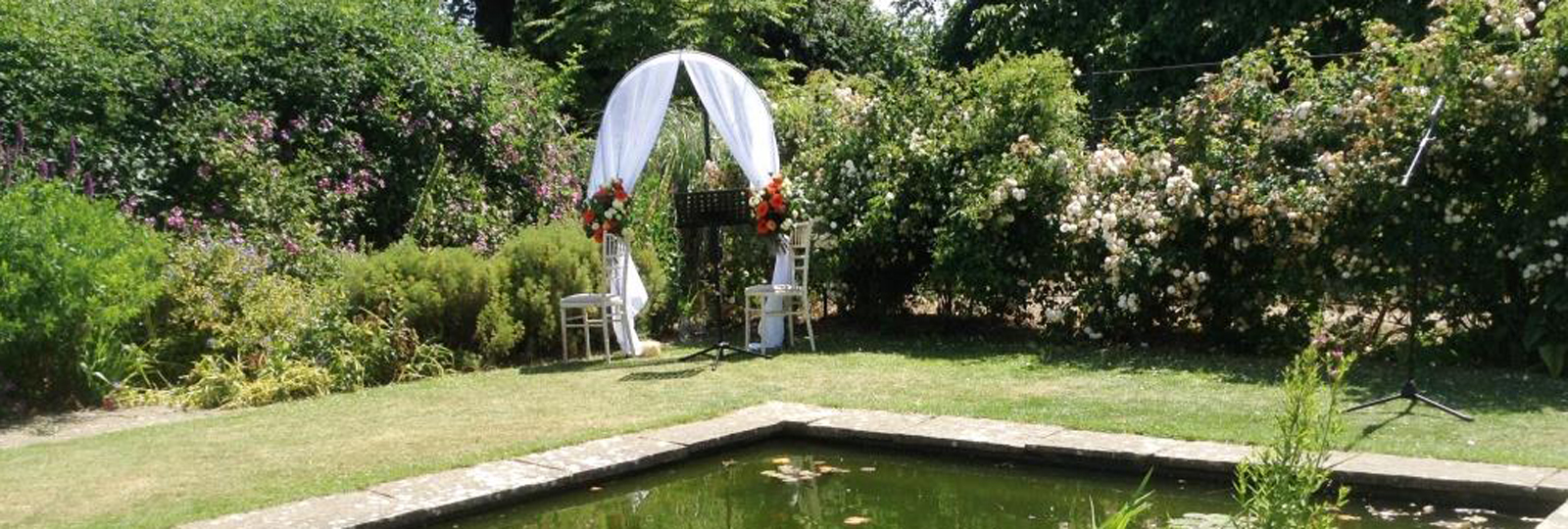 A wedding ceremony at Preston Manor Gardens. a corner of the walled garden is decorated with an archway draped with white curtains and flowers ready for a wedding ceremony. A large pond is in the foreground.