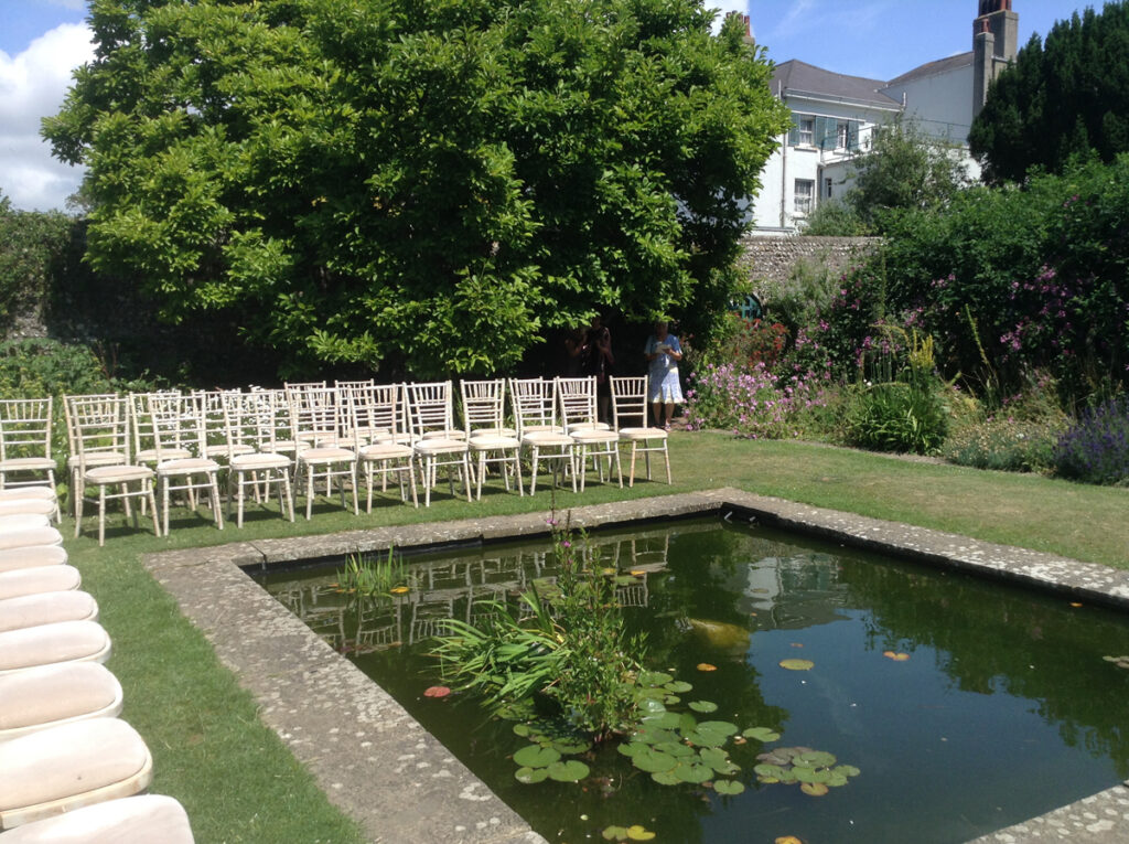 A wedding ceremony at Preston Manor Gardens. A walled garden with a large square pond, with fish and lilies. Rows of chairs are along two side of the pond facing the corner of the garden