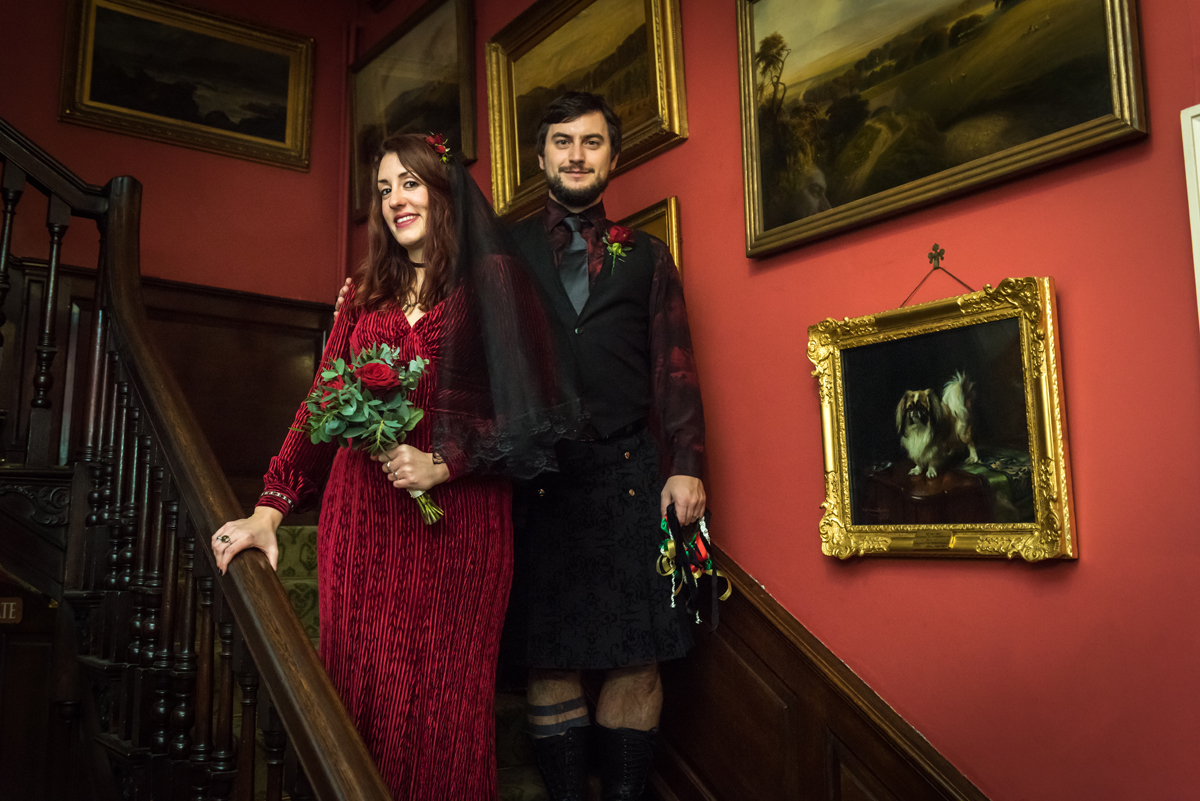 A wedding couple at Preston Manor. A man and a woman stand on the stairs inside the Manor, she is holding a bouquet of flowers and wears a red dress, he wears a kilt.
