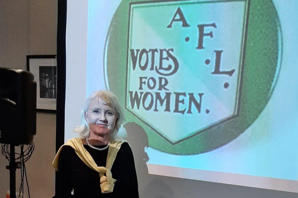 Louise Peskett stands by a screen with an image of a shield and the words A.F.L Votes for Women