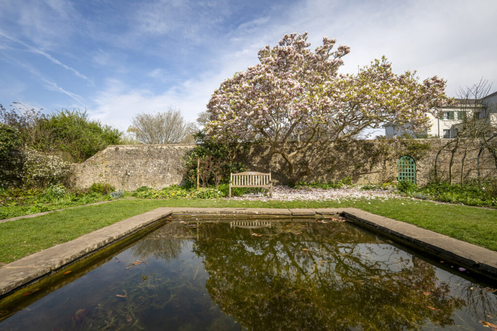 Preston Manor Gardens. A large rectangular pond is in the foreground of a walled garden. A wooden bench and tree are reflected in the pond distance.