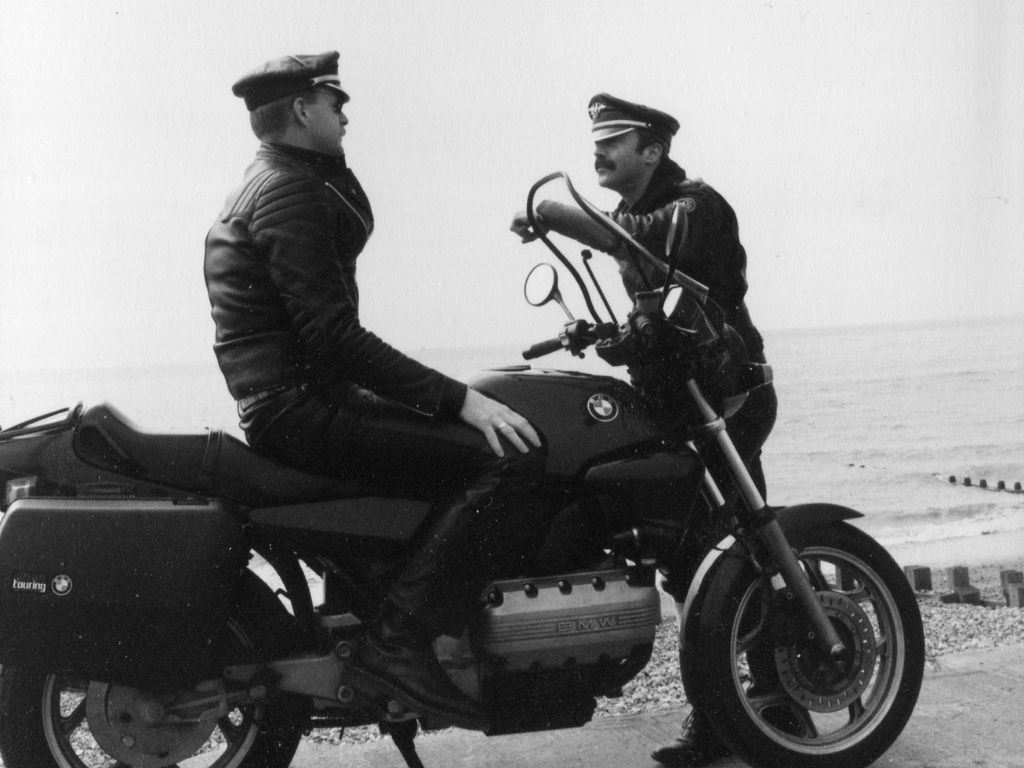 Sussex Lancers. Two men wearing leather jackets, trousers, and hats, one is sitting on a BMW motorbike while the other stands in front of it.