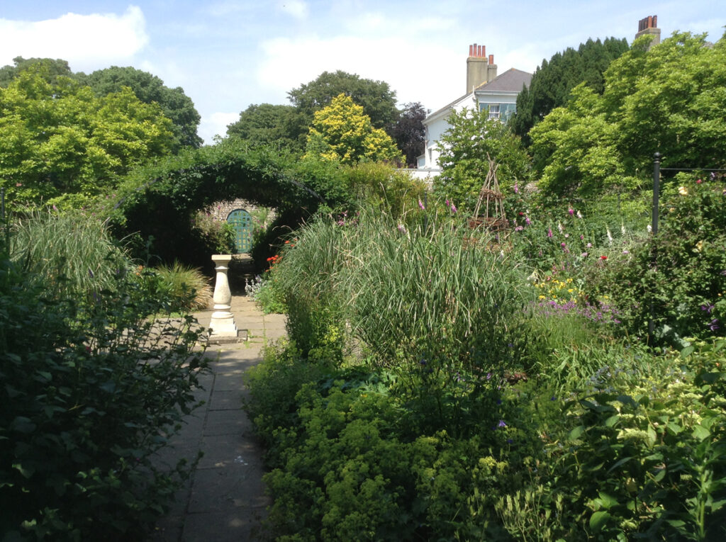 Preston Manor Gardens with a view towards kitchen gardens. A path winds down the centre of a bushy garden. A sundial is in the centre of the path and the top of the manor can be seen through the trees to the right.