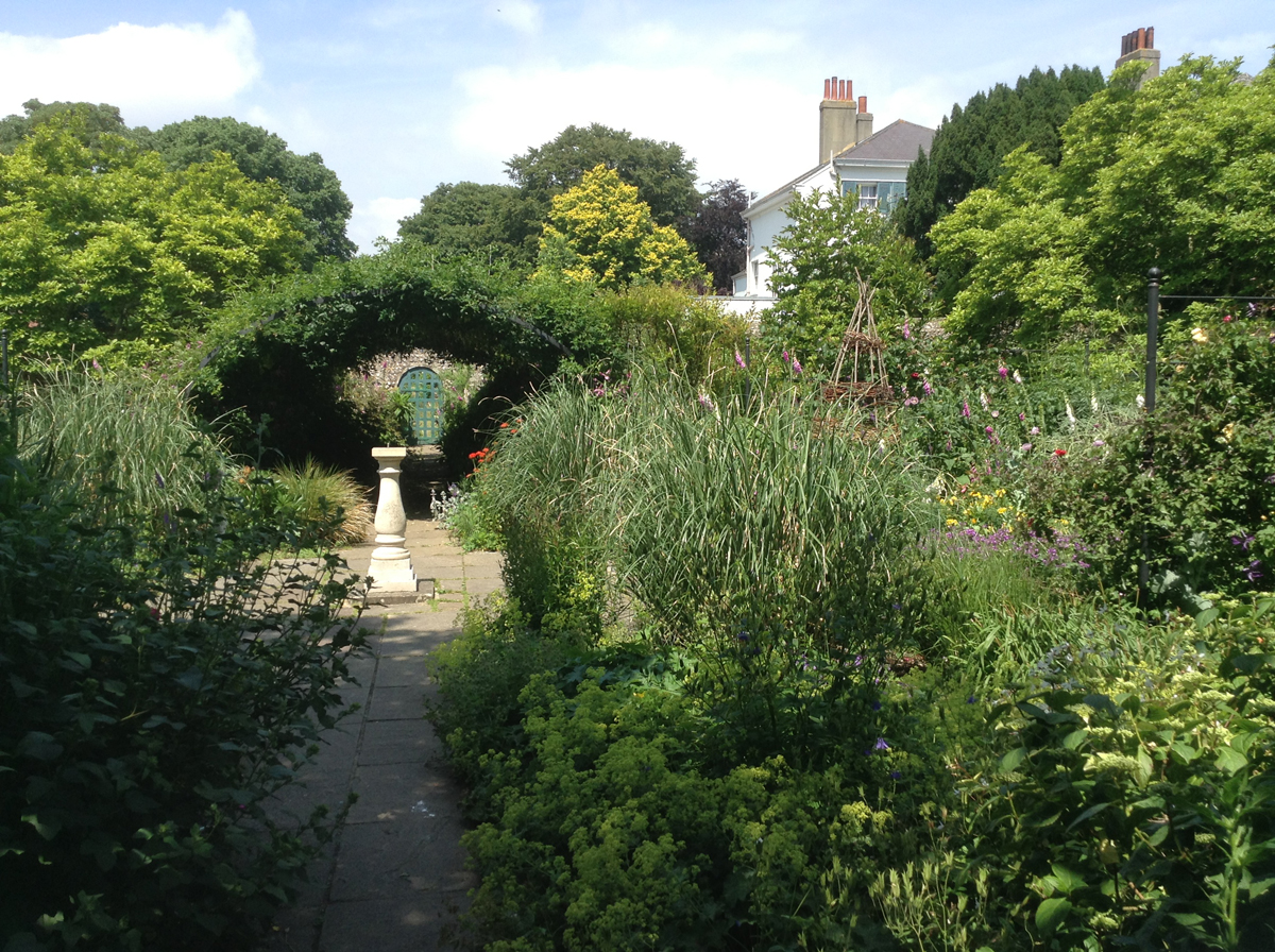 Preston Manor Gardens with a view towards kitchen gardens. A path winds down the centre of a bushy garden. A sundial is in the centre of the path and the top of the manor can be seen through the trees to the right.