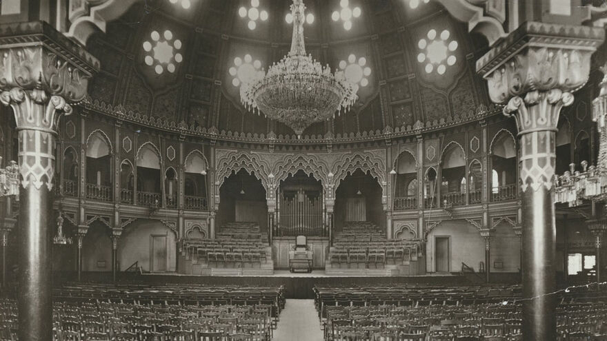 Brighton Dome Chandelier. An ornate round concert hall is filled with chairs facing the stage. A large chandelier hangs overhead