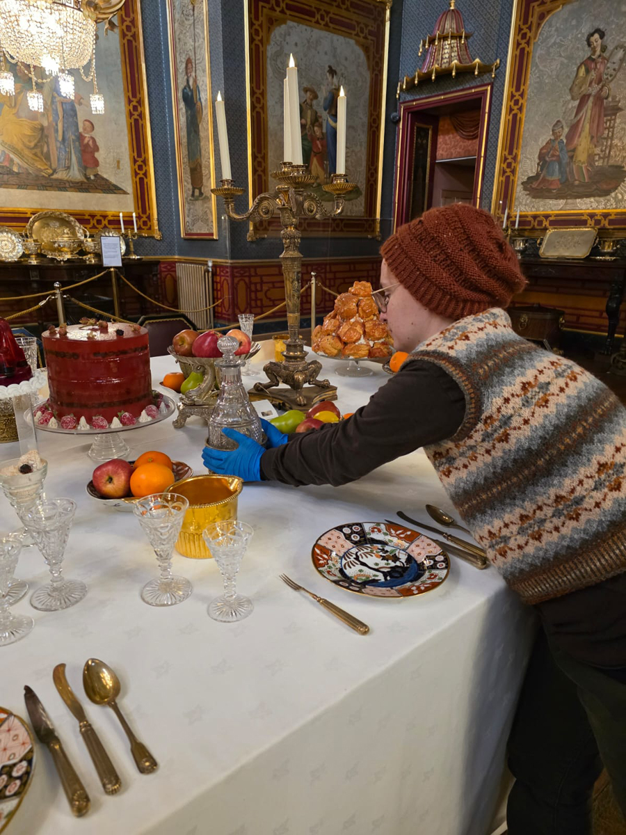 Helping to install the Christmas feast on the Banqueting Room Table. A member of Conservation is leaning over the banqueting table. Wearing gloves, they adjust the table display