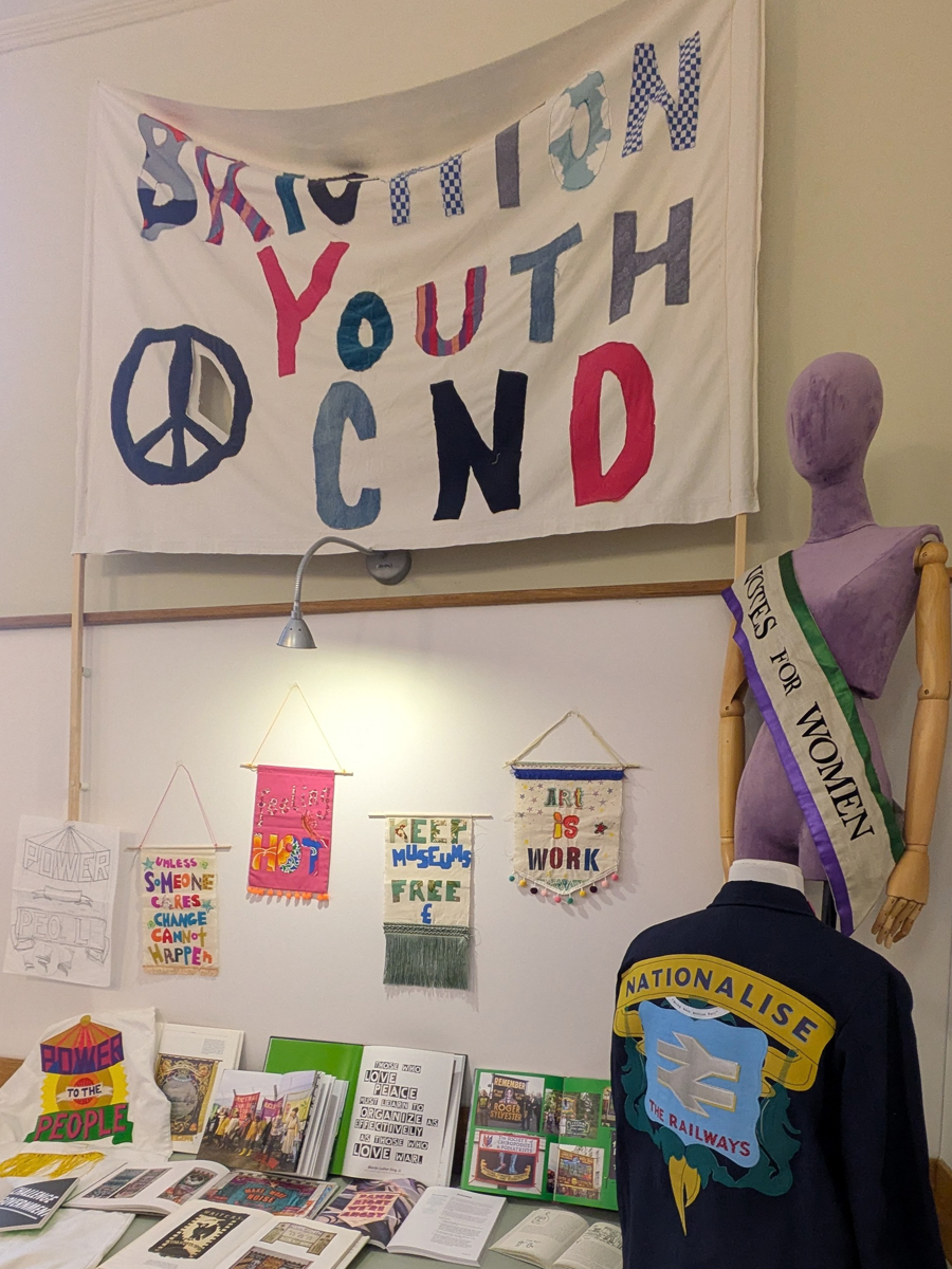 Power to the People session. Books are open on a table, handmade banners are pinned to the wall behind it and two mannequins are to the right. All have messages of protest