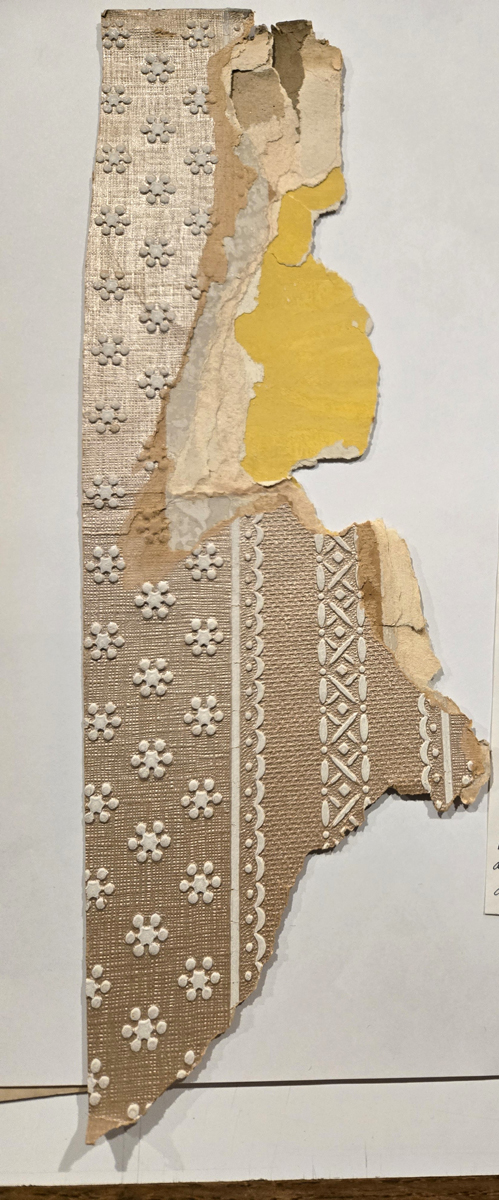 Wallpaper traced to Preston Manor. The torn strip of wallpaper reveals many layers beneath it. The top design is a darkened cream with white flowers and line patterns.