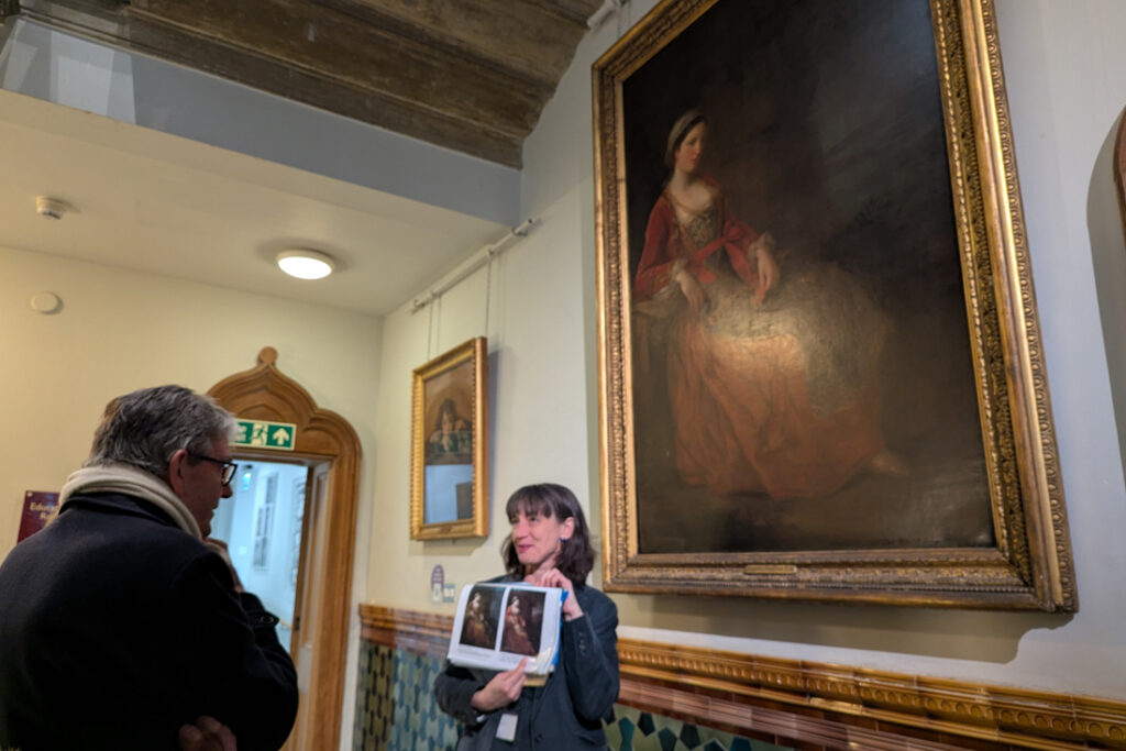 Curator at Work - Alexandra Loske stands in front of a painting in Brighton Museum and talks to a group. She holds a folder of art images to accompany her talk