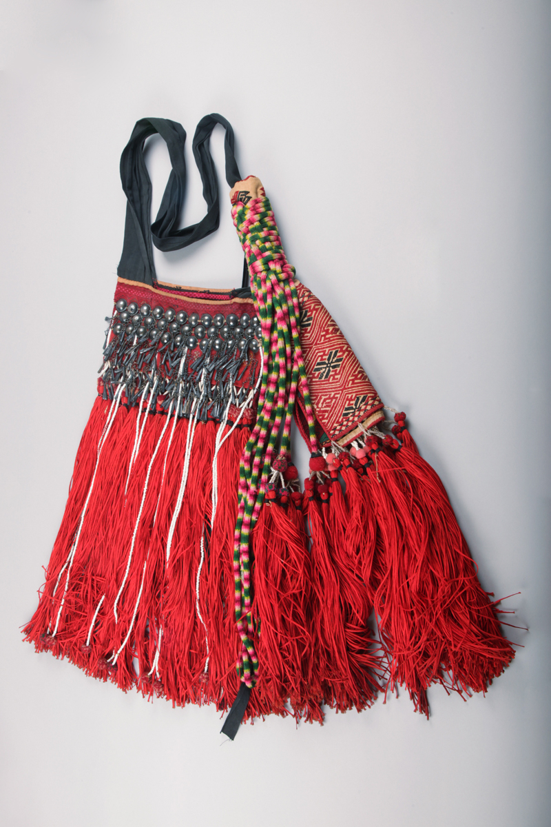 A red and black woven cotton bag from Burma (Myanmar) associated with the Kachin cultural group. It is decorated with pink, green and yellow brocaded diamonds patterns. It has woven cotton attached to strap and also with white beads and red cotton tassels. It was collected in Kachin State in 1920s.