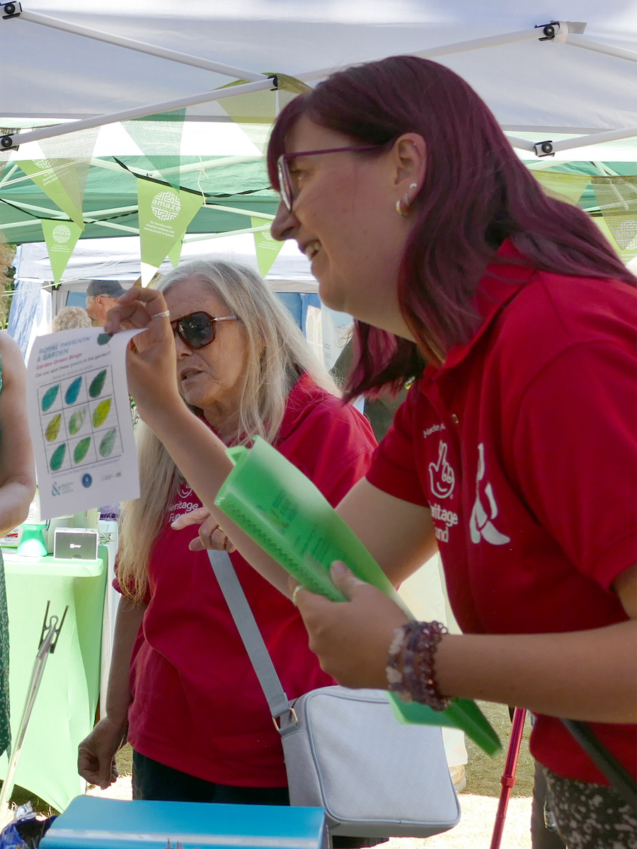 Royal Pavilion Garden Greeters 2026. Two women in red polo shirts with the HLF and B&HM logos talk to visitors out of shot. The woman in the foreground is holding a green plastic wallet of handouts in one hand, and a small leaflet with different shades of green in the other.