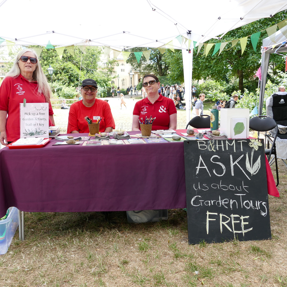 Royal Pavilion Garden Greeters 2026. Three women in red polo shirts with the HLF and B&HM logos. Two are seated and one is standing. They are all behind a table covered with a purple tablecloth and leaflets, signs and pots of pencils. A large chalkboard is propped up in front of the table. It reads B&HMT ask us about Garden Tours Free.