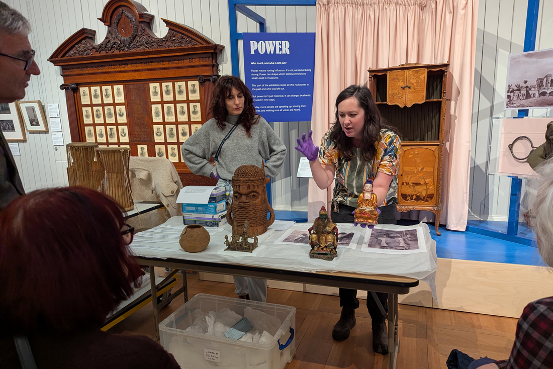 Curator at Work - Portia Tremlett. Portia stands behind a table in the Central Gallery of Brighton Museum. Objects from the World Art collection are on the table and Portia holds a Chinese Deity object as she speaks