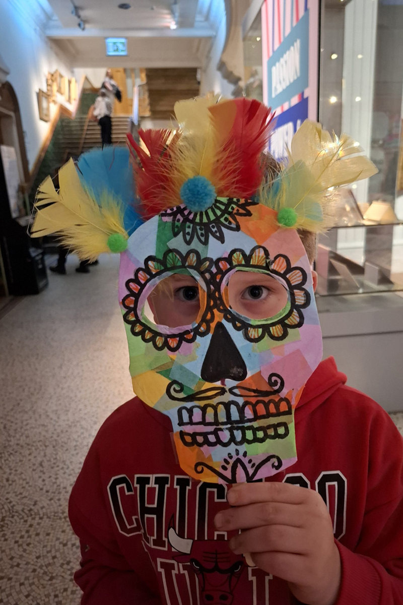 Art Club at Brighton Museum. A boy holds a mask to his face. The mask is a skull with feathers attached to the top.