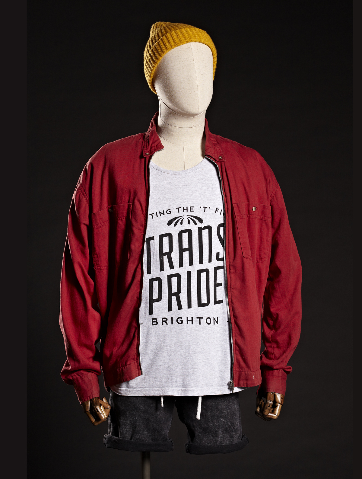 An outfit for the Queerlooks exhibition. A male mannequin wears a yellow wool hat, red jacket, white t-shirt with "Trans Pride" wording and black shorts. Credit Tessa Hallmann.