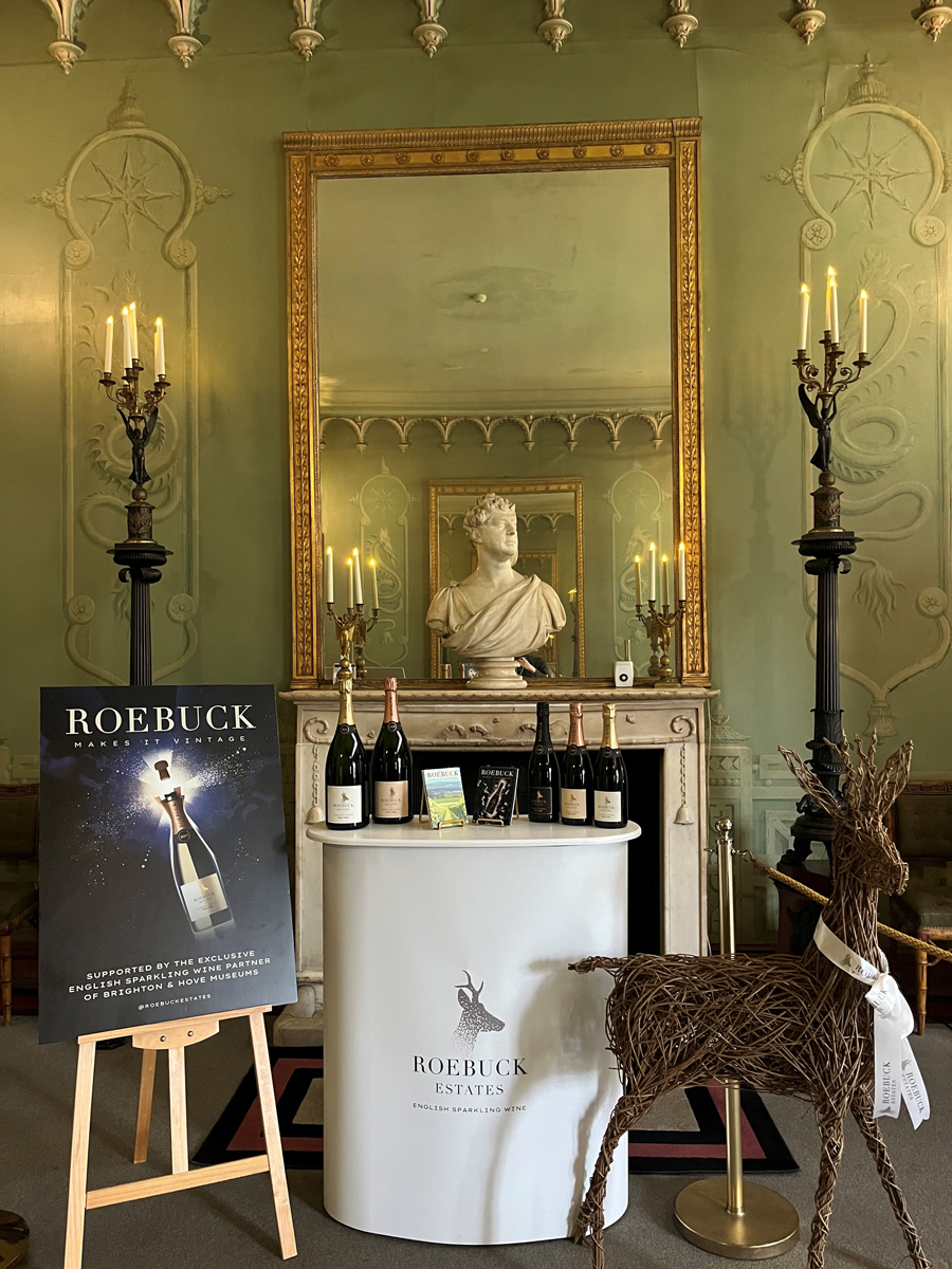 Roebuck Estates wine promotional image inside the Royal Pavilion. A temporary bar plinth with bottles of wine is placed in front of a fireplace. A bust of King George IV sits on the mantle above. To the left of the bar is a Roebuck Wine poster on a wooden easel. A wicker sculpture of a deer - the Roebuck logo - is placed to the right of the bar.