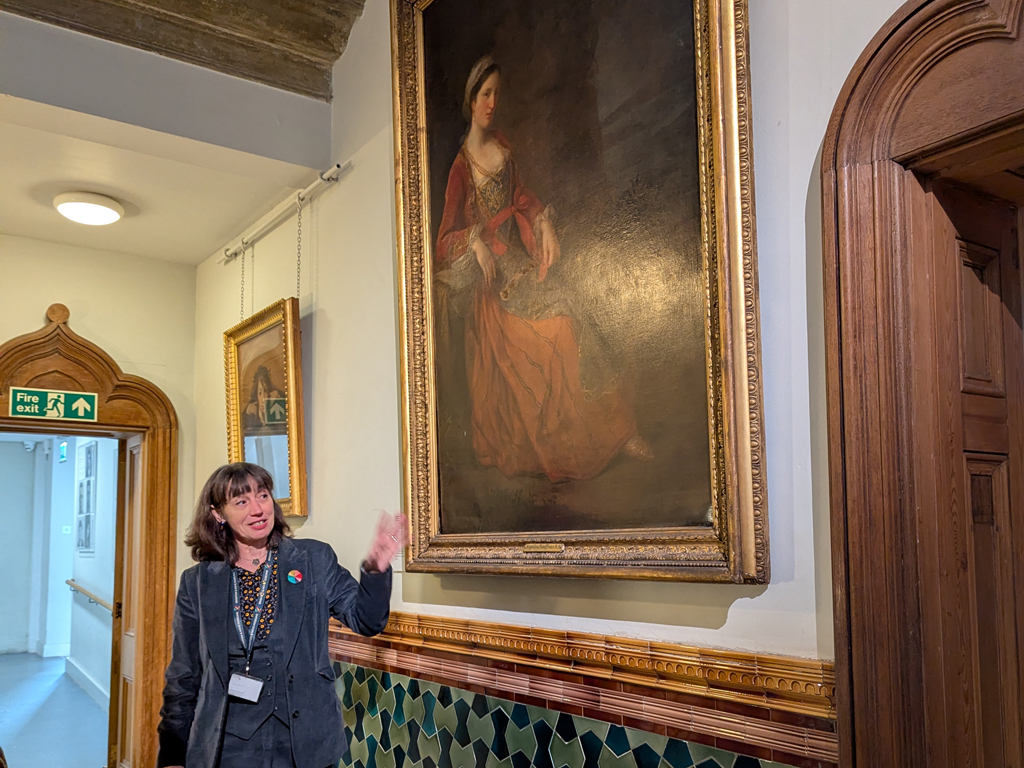 Curator at Work - Alexandra Loske stands in front of a painting in Brighton Museum and talks to a group. She holds a folder of art images to accompany her talk