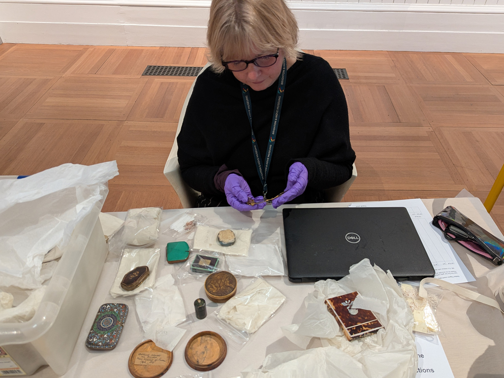 Curator at Work - Lucy Faithful. Lucy sits behind a desk in the Central Gallery of Brighton Museum. On the desk is a laptop and a plastic storage box containing various wrapped objects. A group of people stand facing the desk at Lucy unwraps and talks about the objects.