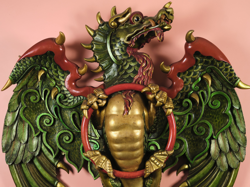 Dragon currently outside William IV Room of The Royal Pavilion. A 3d relief of a green dragon with red tips on its wings and gold claws and belly. It holds a red hoop and looks away to the right.