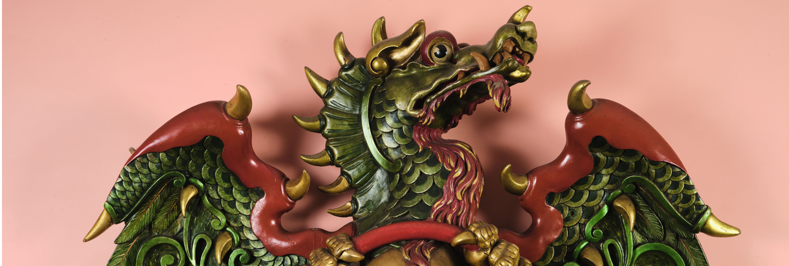 Dragon currently outside William IV Room of The Royal Pavilion. A 3d relief of a green dragon with red tips on its wings and gold claws and belly. It holds a red hoop and looks away to the right.