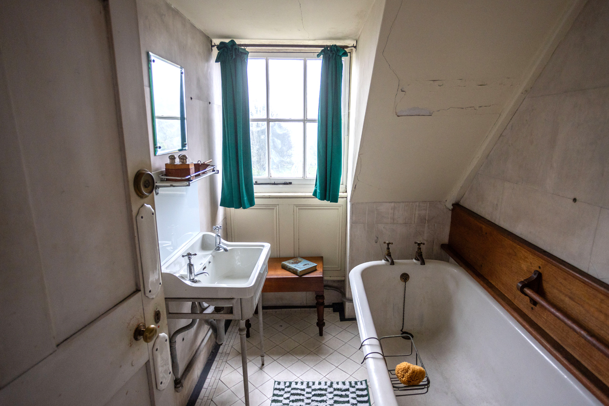 Preston Manor Bathroom. A white door is open on the left to reveal a bath on the right, sink on the left and a window with green curtains on the far wall