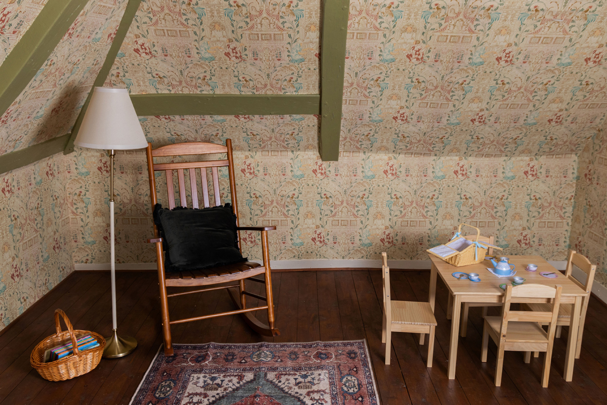 The Nursery at Preston Manor. A rocking chair is placed beside a standing lamp and basket of books to the left of the room. A child's square wooden table and 4 chairs are to the right. A tea playset and basket are on the table